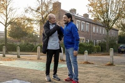 Twee mannen staan buiten in een woonwijk en lachen terwijl ze met elkaar praten. Bomen op de achtergrond.
