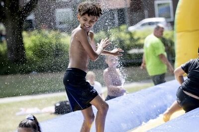 Een jongen met krullend haar speelt op een natte glijbaan, omringd door andere kinderen in een zomerse setting.